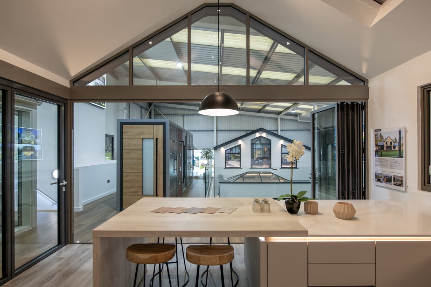 Modern kitchen with a high vaulted ceiling, large glass windows, a light wooden island with two stools, a hanging black lamp, and decorative items including a white orchid. Sliding doors and glass walls offer views of adjoining rooms.