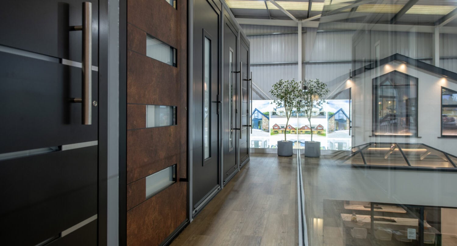 Modern indoor showroom with various large doors, including bifold doors, on display. Wood flooring, a glass railing, a potted plant, and bright natural light streaming through large windows in the background.