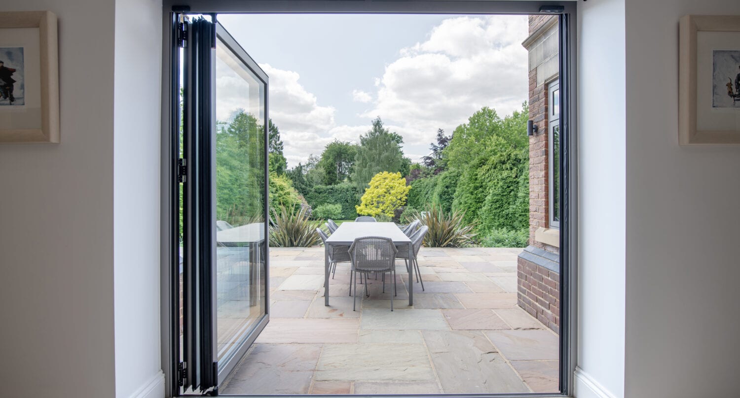 Open glass patio doors lead to a stone terrace with a table and chairs, surrounded by lush green trees and plants under a partly cloudy sky.