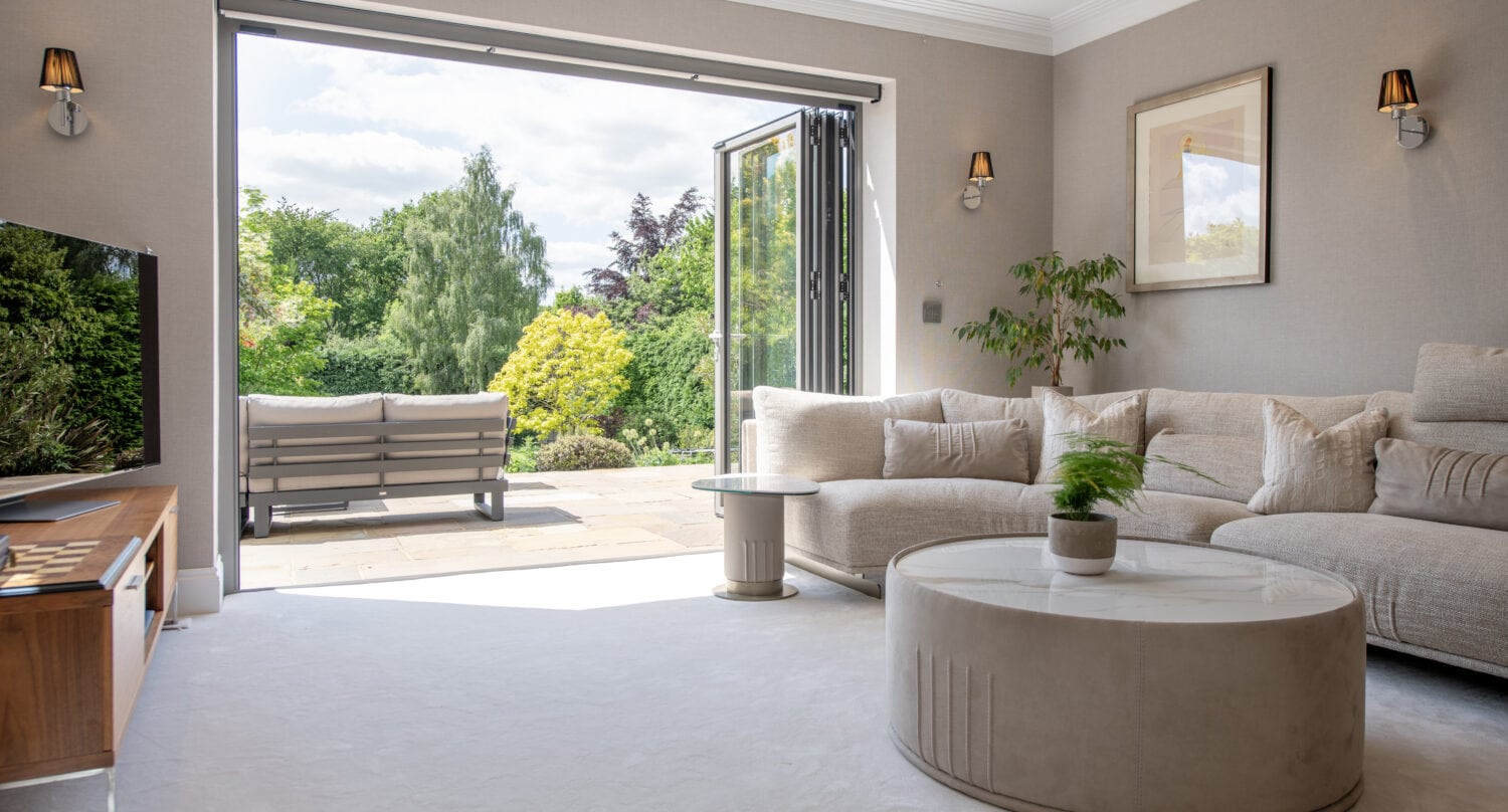 A bright living room with a beige sectional sofa, round coffee table, TV, and wall art. Glass doors open to a patio with outdoor seating, overlooking a lush garden with trees and bushes.