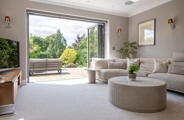 A bright, modern living room with a beige sectional sofa, round coffee table, and wall-mounted TV. Glass doors open to a patio with outdoor seating and a lush green garden view.