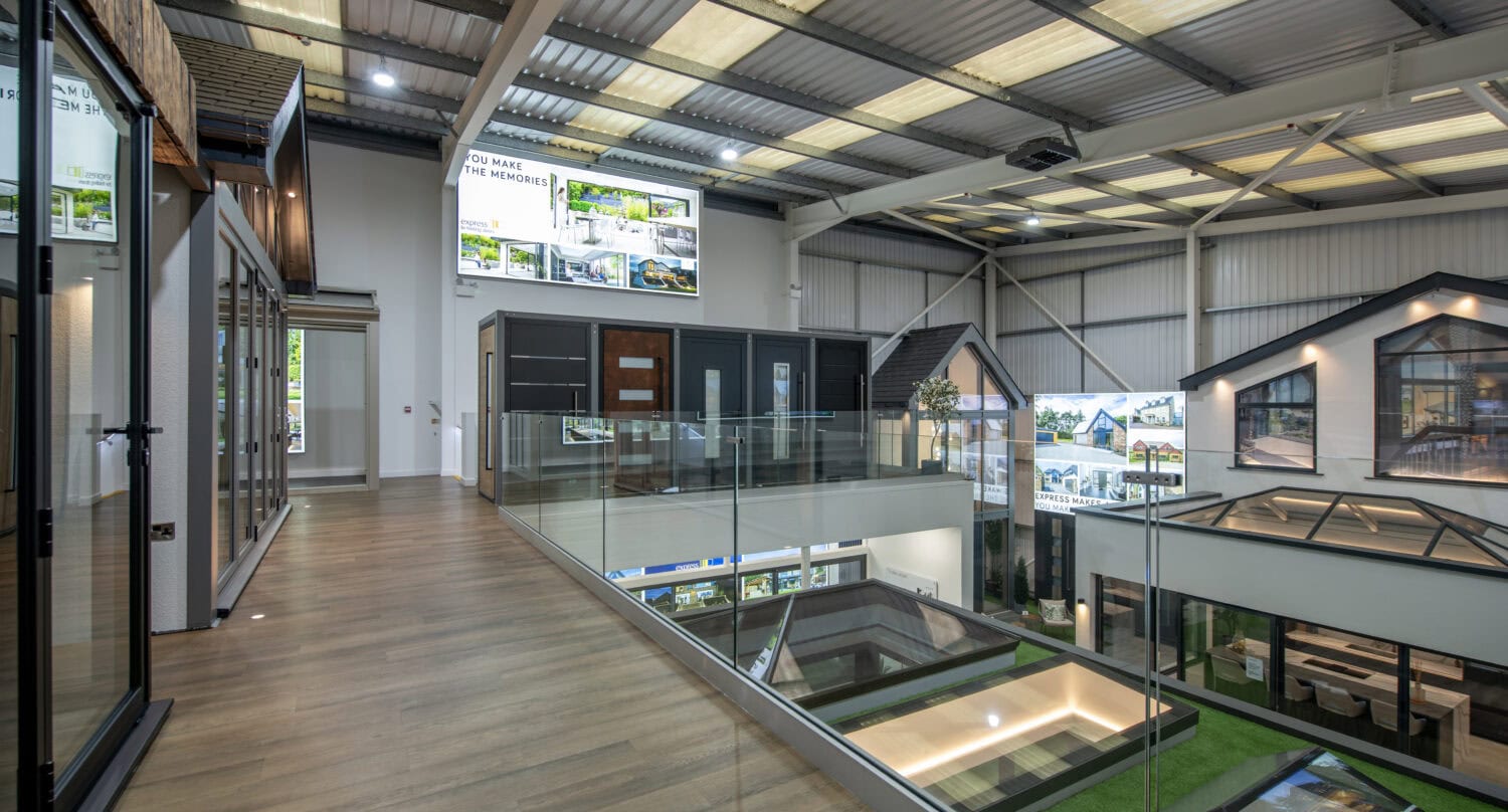 Interior of a modern showroom featuring sample house facades, large windows, bifold doors, glass railings, wood flooring, and illuminated display signs under a metal ceiling.