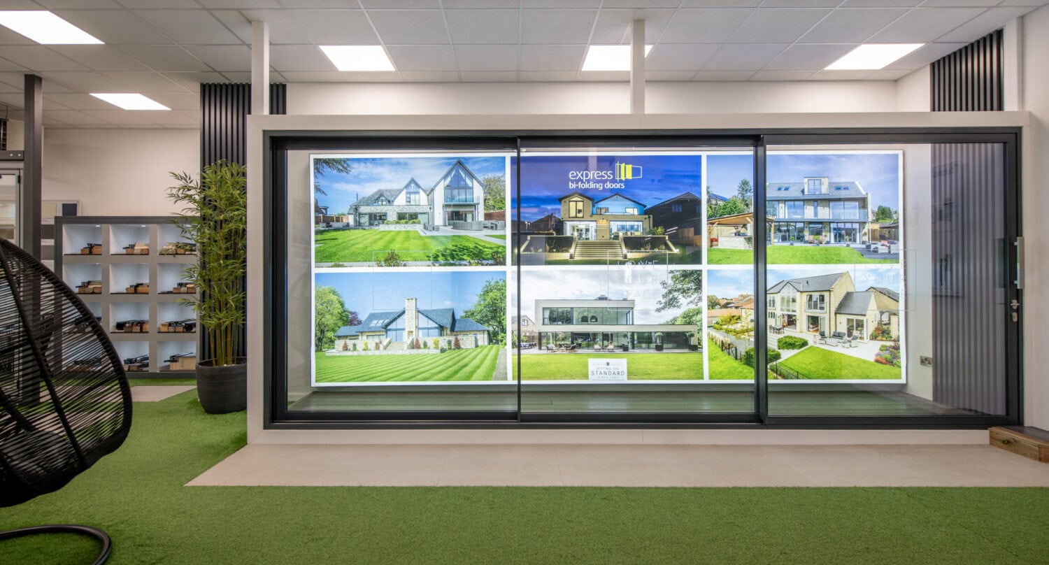 A modern real estate office features a large illuminated display wall with photos of different houses. The space has artificial green grass flooring, bespoke glass solutions, shelves, and contemporary decor.