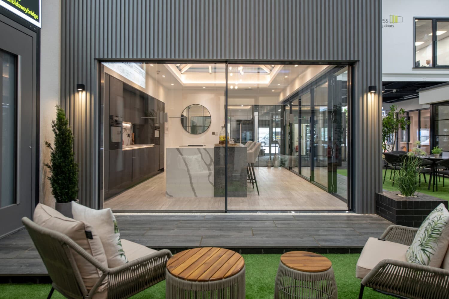 Modern indoor kitchen and dining area viewed through bespoke glass solutions with large sliding doors, opening onto a cozy outdoor seating area with chairs, a small table, and lush greenery in the foreground.