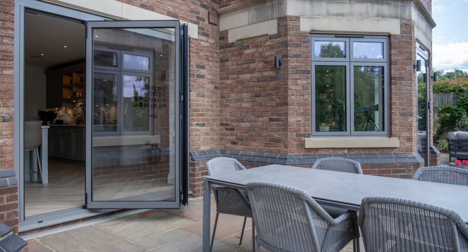 A folding glass door opens from a brick house onto a patio with a gray outdoor dining table and six woven chairs. The patio is paved with stone tiles, and there are trees visible through the window.