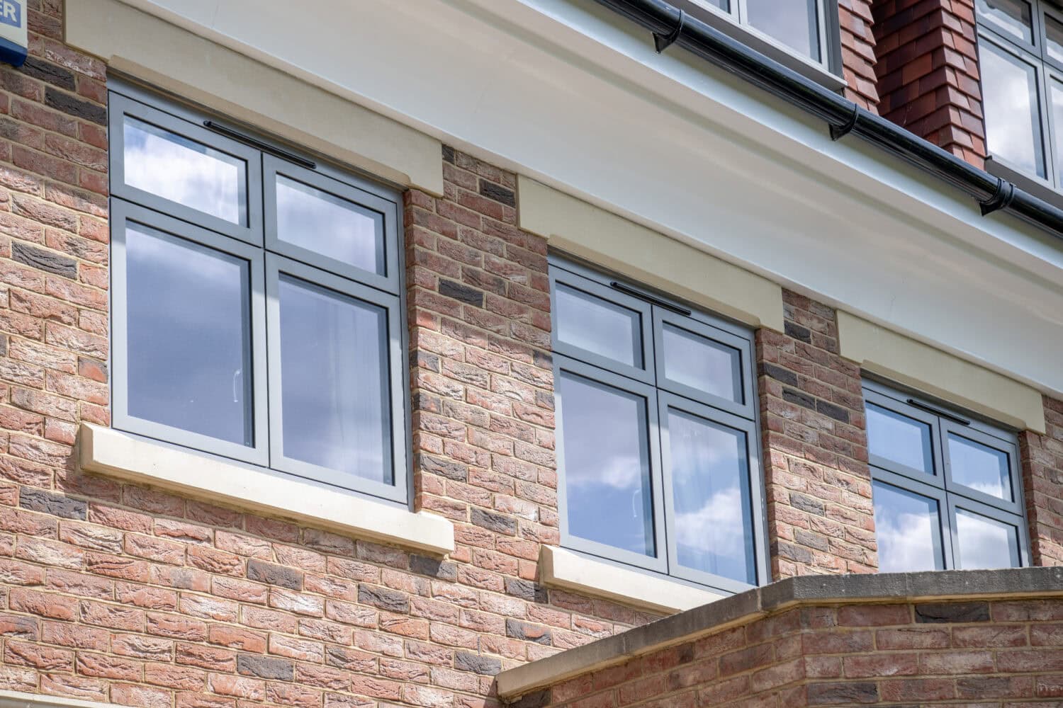 Close-up of two large, modern double-glazed windows on a brick building, reflecting a blue sky with clouds. The windows have gray frames and are bordered by light-colored stone accents.