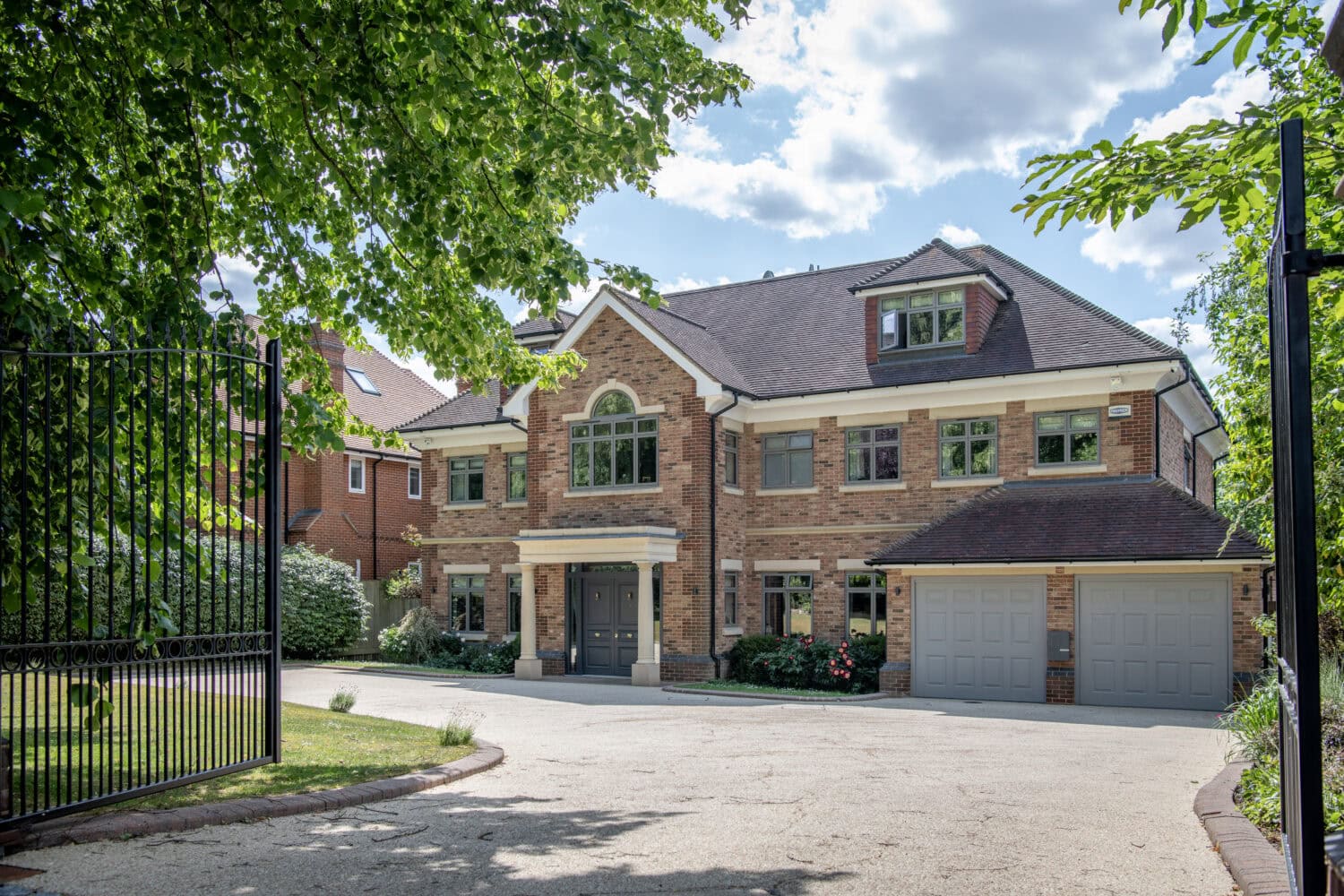 A large, modern brick house with a symmetrical facade, tall windows, and double garage, surrounded by greenery and partially shaded by trees, viewed from behind an open black metal gate on a sunny day.