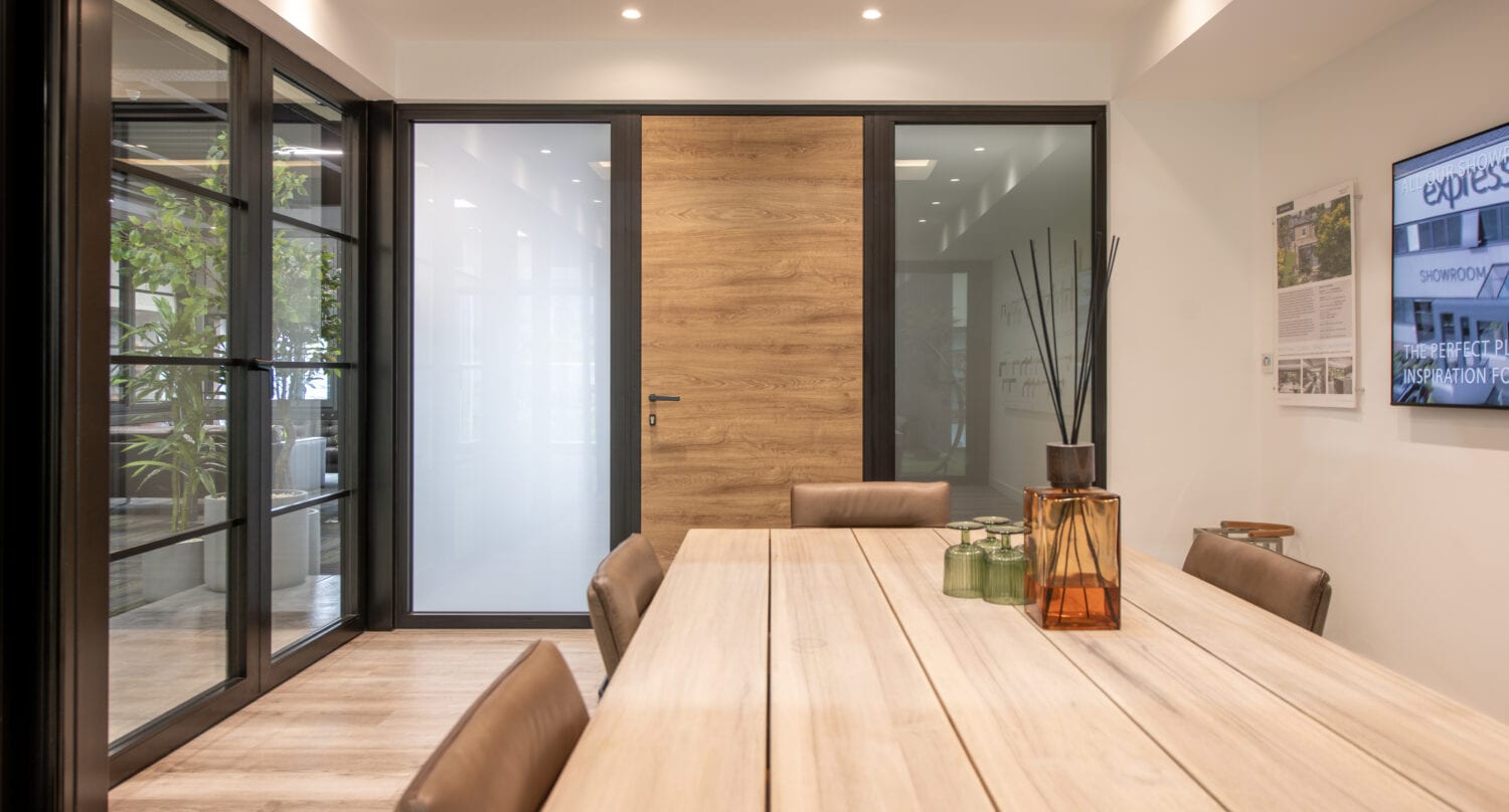 Modern conference room with a light wooden table, brown leather chairs, and decorative vases. Bespoke glass solutions are showcased in the glass doors, while a large wooden front door and wall-mounted screen complete the space, lit by a skylight above.