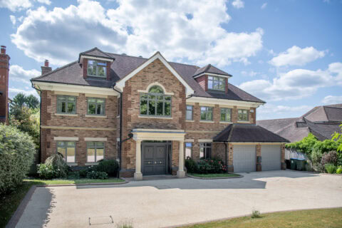 A large, modern brick house with three stories, arched windows, a double front door, and an attached two-car garage. The driveway is spacious, surrounded by greenery, under a partly cloudy sky.