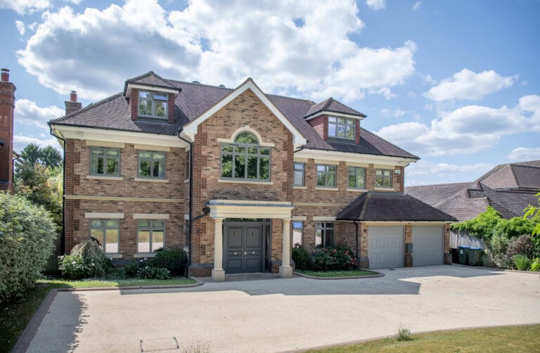 A large, modern brick house with three stories, arched windows, a double front door, and an attached two-car garage. The driveway is spacious, surrounded by greenery, under a partly cloudy sky.
