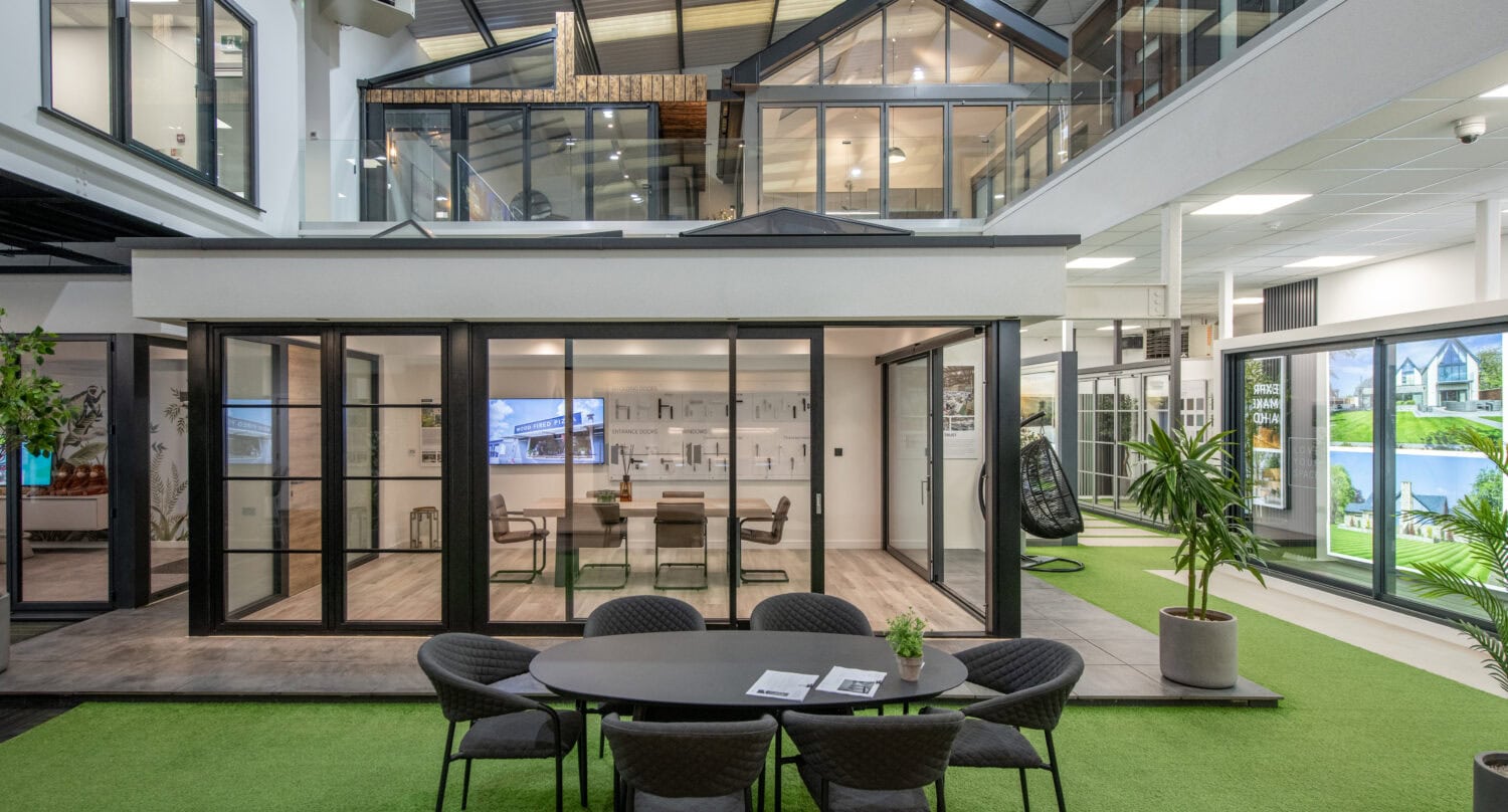 Modern indoor showroom with artificial grass flooring, featuring a round table and chairs in the foreground. Glass-walled office spaces and display rooms showcase bespoke glass solutions like bifold doors in the background, resembling small houses.